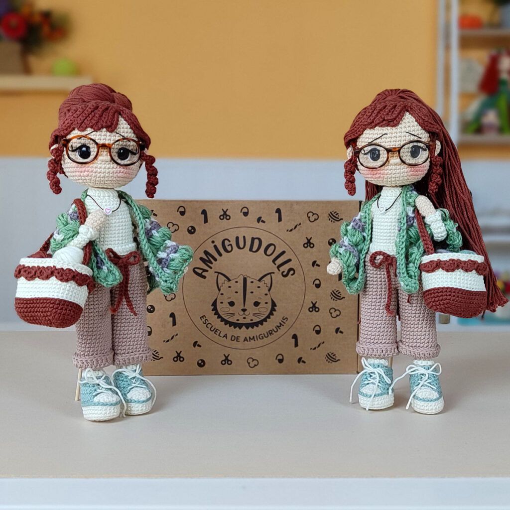 Muñeca amigurumi Nora Merino, con chal verde y morado, bolso y zapatillas, junto a la caja del kit de materiales Amigudolls.