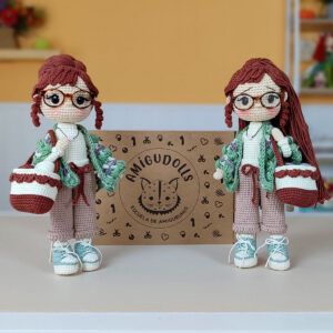 Muñeca amigurumi Nora Merino, con chal verde y morado, bolso y zapatillas, junto a la caja del kit de materiales Amigudolls.