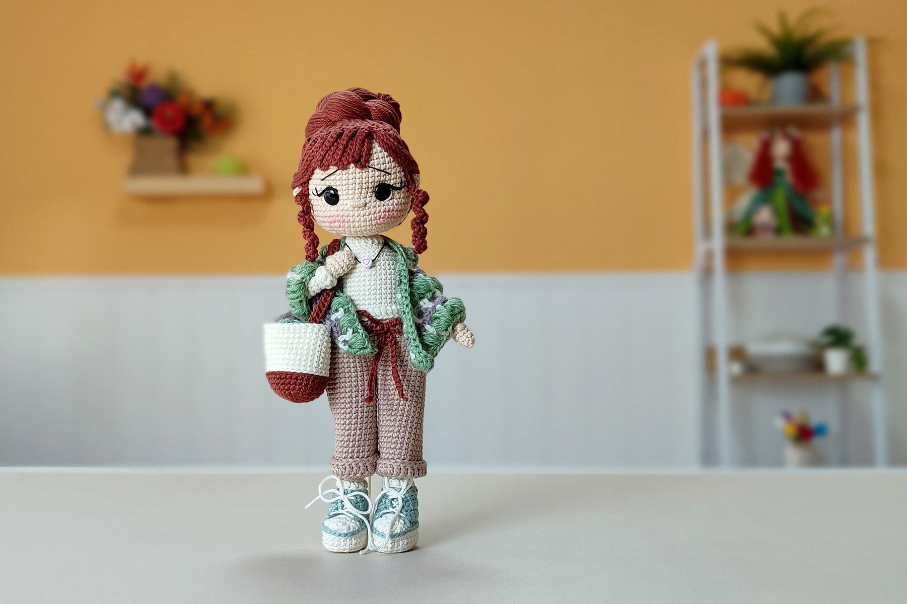 Muñeca amigurumi Nora Merino con chal verde y morado, bolso con ovillos y zapatillas, sobre un fondo mostaza.